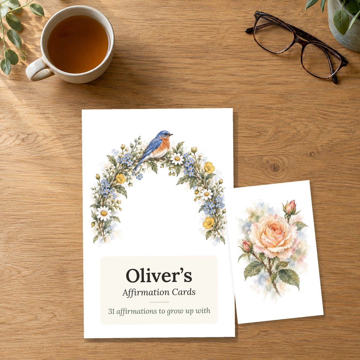 The Oliver's Affirmation Cards box front beside a card with a watercolor-style flower illustration, on an oak table
